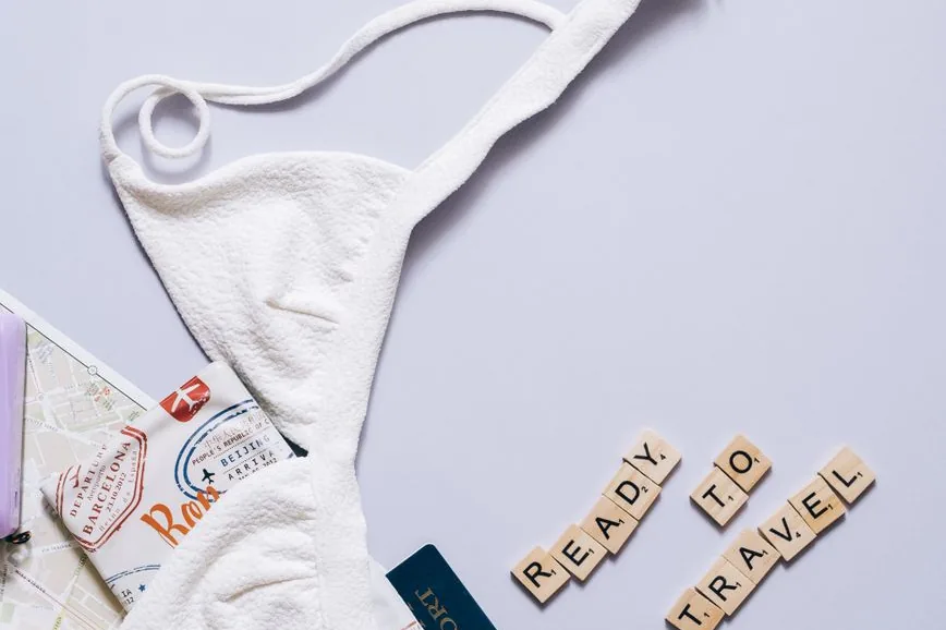A white bikini top, a map, a passport and Scrabble tiles spelling READY TO TRAVEL on a pale surface