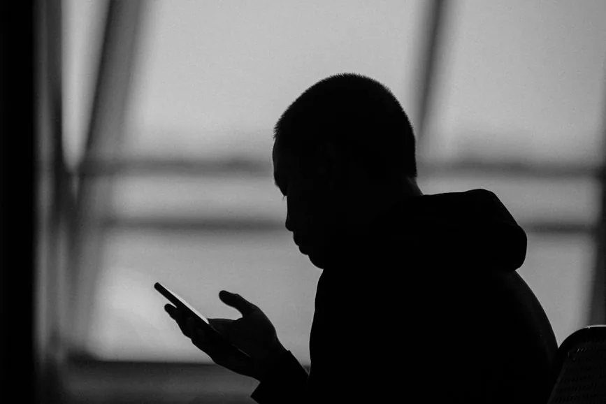 A black-and-white silhouette of a person looking at a phone in front of a large window