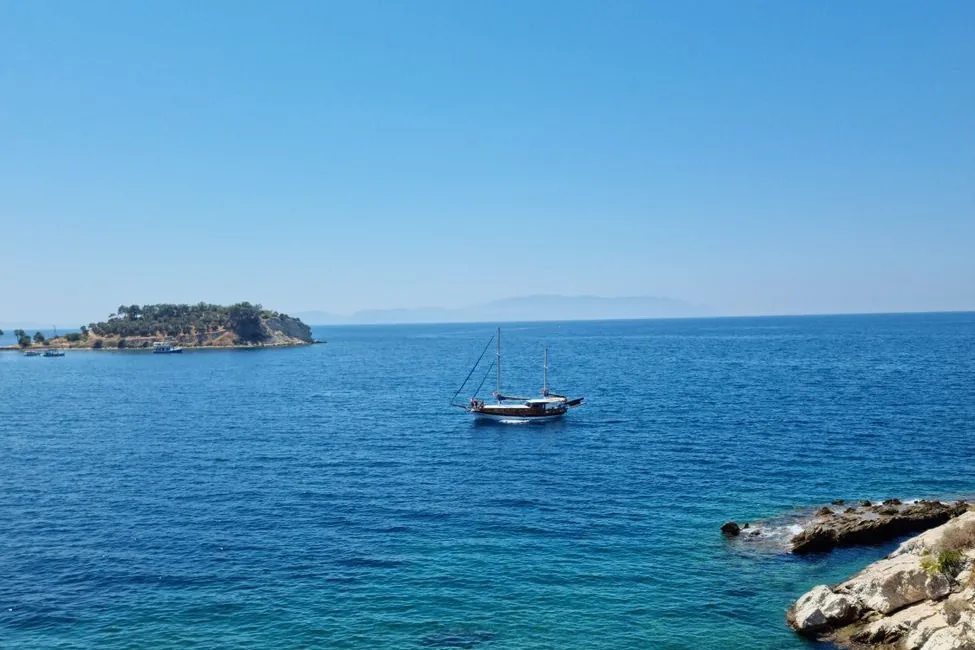A two-masted wooden sailing boat on calm blue sea near a small wooded island