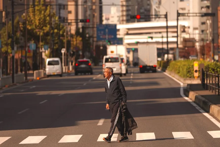 An older man walking across a zebra crossing on a wide street in Japan, with cars and vans behind