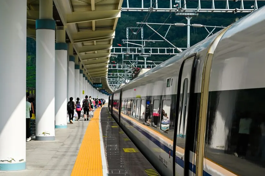 A white high-speed train with a dark window band and a blue stripe at a platform, passengers walking beside it