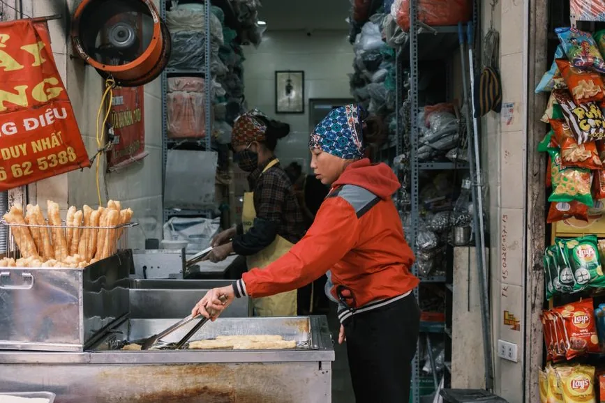 A woman frying dough sticks at a street-food stall in Vietnam, with bags of snacks hanging by the door